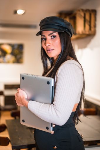 A vertical shot of a young Latina holding a laptop in a cafe while looking directly into the camera