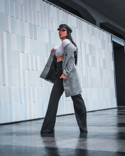 A stylish woman with a classy leather hat and a gray coat posing in front of a gray wall