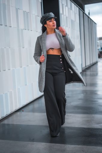 A vertical shot of a woman walking in front of a gray wall in a stylish outfit