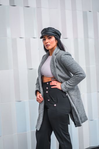 A vertical shot of a young Latina posing in a leather cap and gray blazer in front of a gray wall