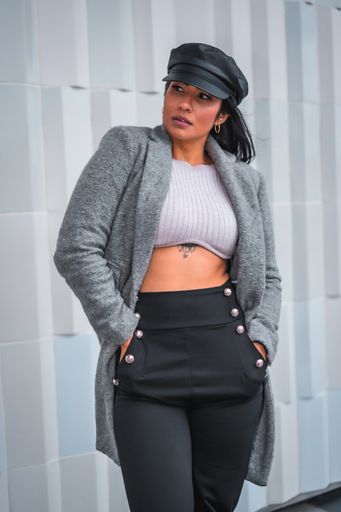 A vertical shot of a woman posing in a classy leather hat and gray open coat in front of a gray wall