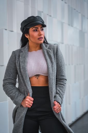 A vertical shot of a woman posing in a leather hat and gray open coat in front of a gray wall