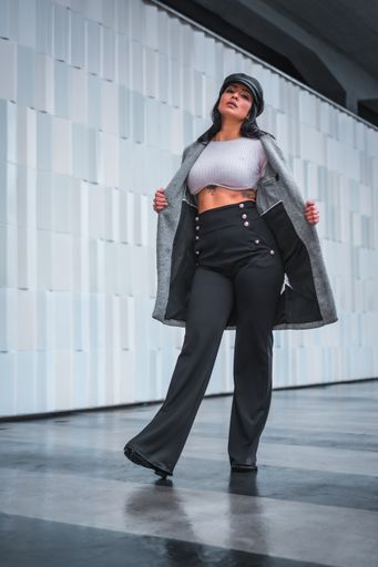 A low angle shot of a woman posing in a leather hat and gray open coat in front of a gray wall