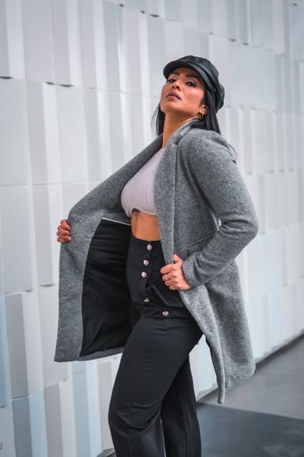 A young woman elegantly posing in a leather hat and gray open coat in front of a gray wall