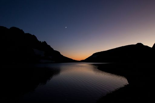 A serene twilight landscape featuring a crescent moon over a calm lake, with silhouetted mountains in the background.