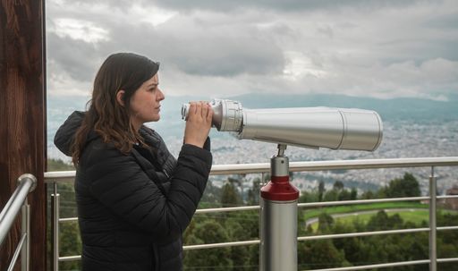Side view of beautiful young woman, standing at a viewing platform, holding in her hands long binoculars mounted on a metal pedestal