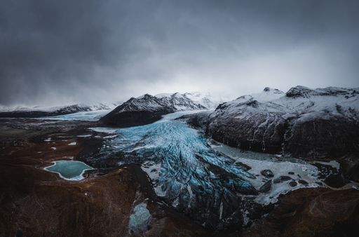 A stunning aerial view of a glacier and surrounding mountains under a dramatic cloudy sky.