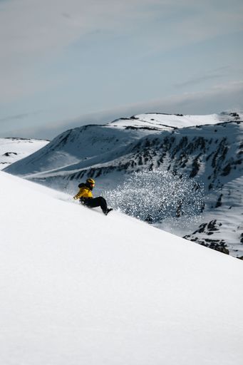 A person in a yellow jacket snowboarding down a snowy mountain slope with a scenic mountain range in the background.