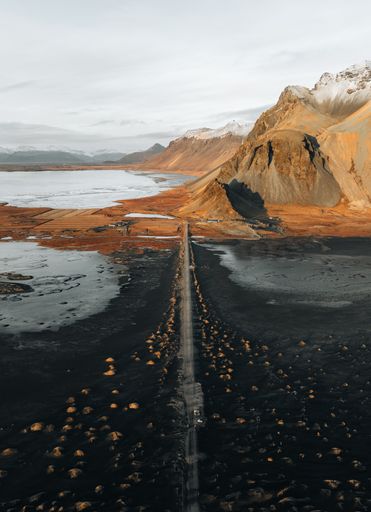 Aerial view of a long road leading through a rugged landscape with mountains and water bodies in the background.