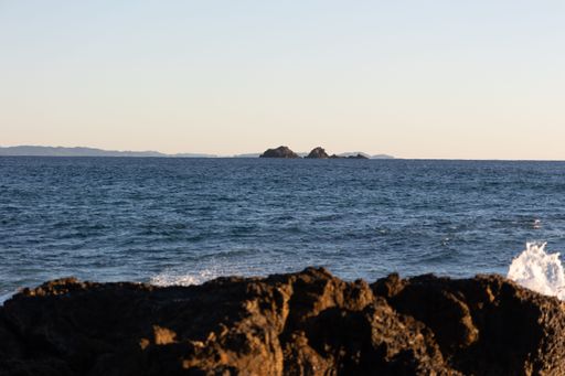 A serene ocean view with distant rocky islands under a clear sky, with waves crashing against the foreground rocks.