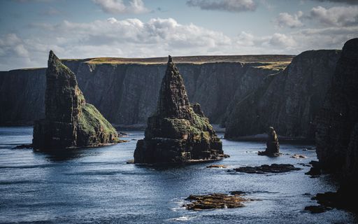 A stunning seascape featuring the Old Man of Hoy sea stack on the Orkney Islands, with dramatic cliffs and blue waters under a partly cloudy sky.