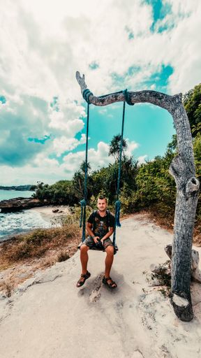A vertical shot of a man sitting on a swing and taking pictures on a beach in Bali, Indonesia