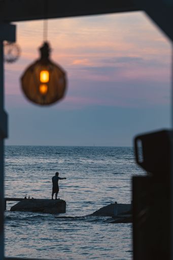 A silhouette of a person fishing on a pier at sunset with a hanging lamp in the foreground.