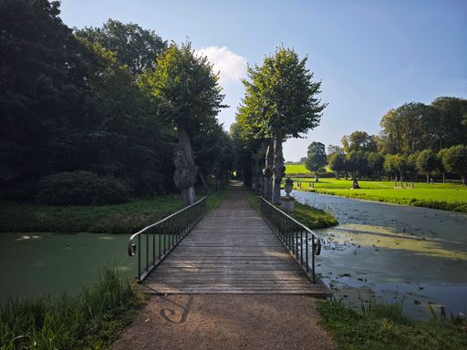 A serene wooden bridge over a tranquil canal surrounded by lush greenery and trees on a sunny day.