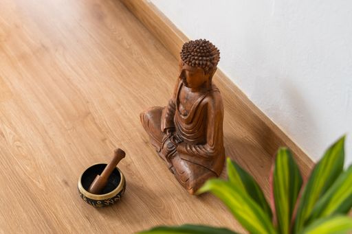 High angle shot of a wooden buddha sculpture next to a Tibetan bowl and plant leaves on a parquet floor.