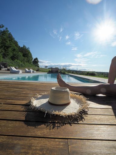 A vertical shot of a woman sunbathing next to a pool with her sunhat next to her, on a sunny day