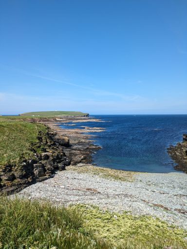 Scenic view of a rocky coastline with clear blue water and a bright sky, featuring grassy cliffs and distant land.