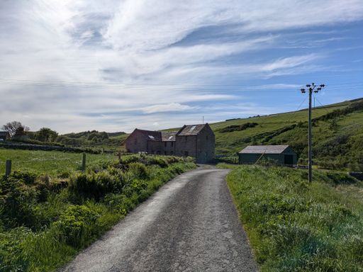 A serene countryside road leading to stone houses surrounded by lush green fields under a partly cloudy sky.