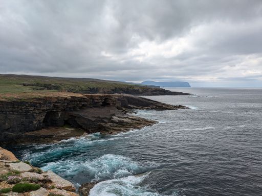 A scenic view of a rocky coastline with waves crashing against the cliffs under a cloudy sky in Orkney, Scotland.