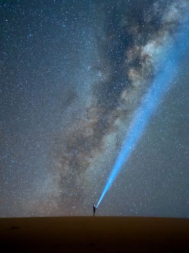 A person standing on a desert hill shining a flashlight into the Milky Way galaxy at night.