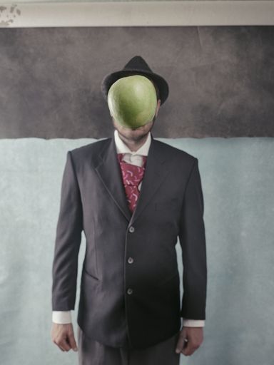 A man in a suit and hat standing near the wall with a green apple in front of his face