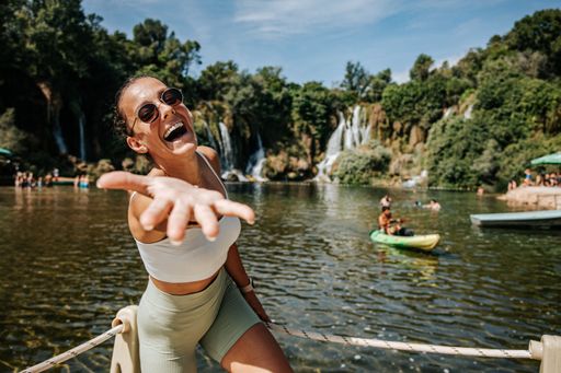 Young woman in sunglasses laughing and reaching out her hand by a scenic waterfall with people kayaking in the background on a sunny day.