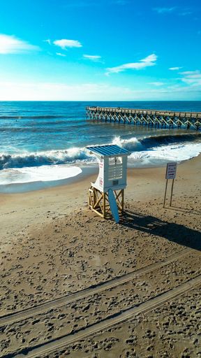 A sunny beach scene with a lifeguard tower, footprints in the sand, and a pier extending into the ocean under a bright blue sky.