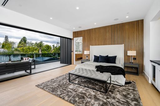 Modern bedroom interior with a large bed, wooden accent wall, and sliding glass doors leading to a balcony with a view of lush greenery.