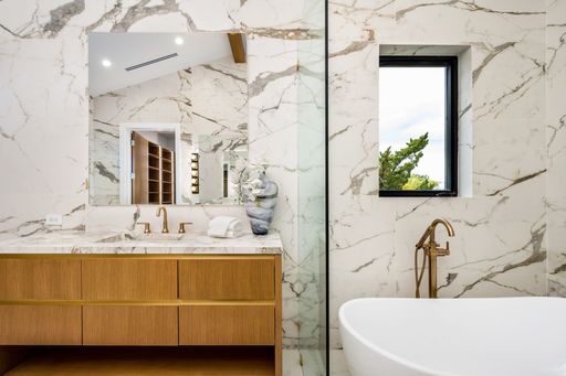 Modern bathroom interior with marble walls, wooden vanity, and gold fixtures, featuring a large mirror and a window with a view of greenery.