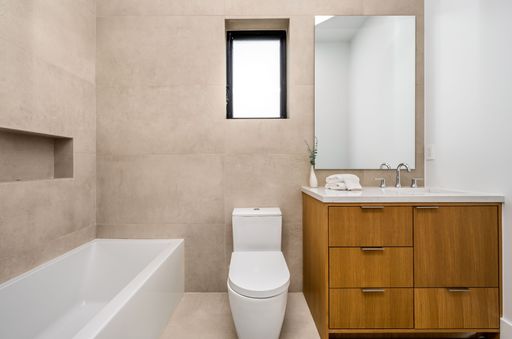 Modern bathroom interior with a bathtub, toilet, wooden vanity, large mirror, and small window, featuring minimalist design and neutral tones.