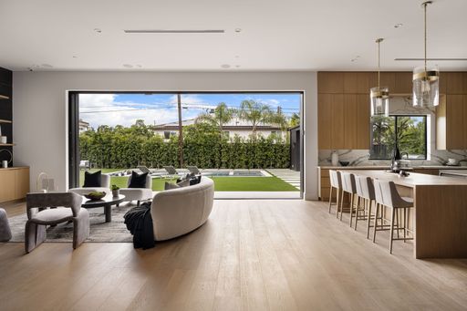 Modern open-concept living room and kitchen with large window overlooking a landscaped backyard with pool and greenery.