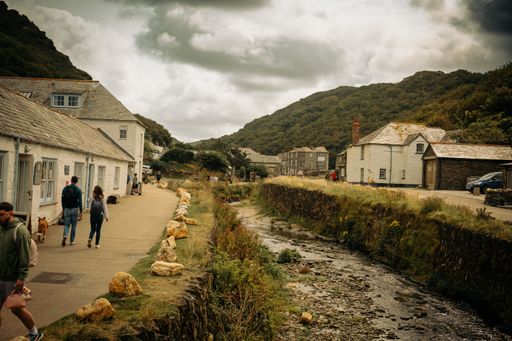 People walking along a charming village path with stone houses and a stream, surrounded by lush green hills under a cloudy sky.