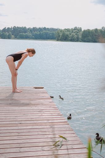 A vertical shot of a Caucasian blonde female in a black swimsuit looking at the ducklings in the lake