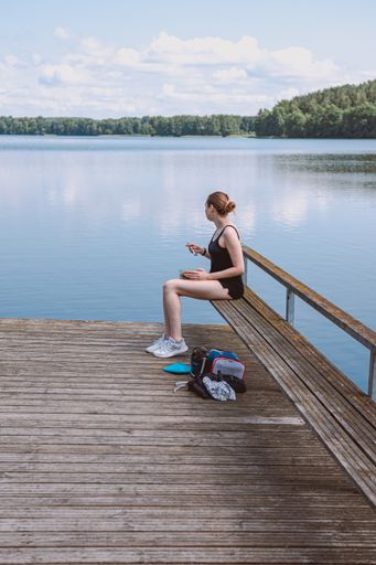 A vertical shot of a Caucasian blonde female in a black swimsuit sitting on the pier in the lake