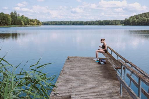 A Caucasian blonde female in a black swimsuit sitting on the pier in the lake