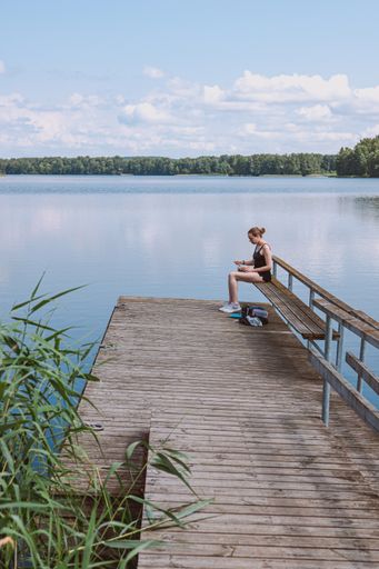 A vertical shot of a Caucasian blonde female in a black swimsuit sitting on the pier in the lake