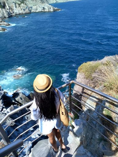 A woman in a hat and white dress standing on a cliffside railing overlooking the ocean.