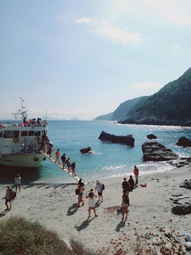 Tourists disembarking from a boat onto a scenic beach with clear blue waters and rocky cliffs under a bright sky.