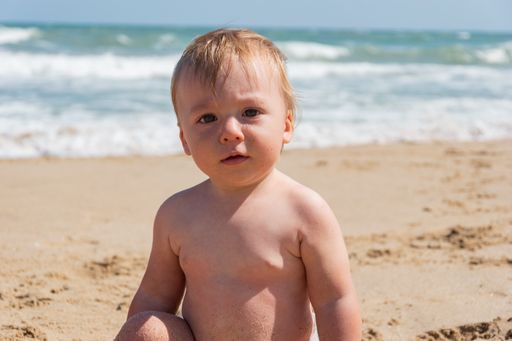 A closeup shot of a cute baby boy sitting on the beach looking at the camera with a curious face