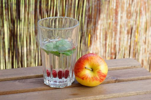 A closeup shot of a glass of water with mint and raspberry with an apple next to it