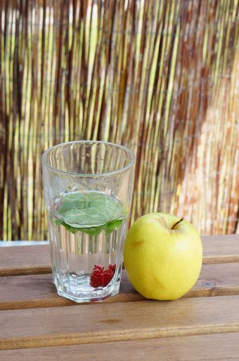 A shallow focus vertical shot of green leaves and raspberry fruit in a glass of water on a wooden table with a yellow apple on the side
