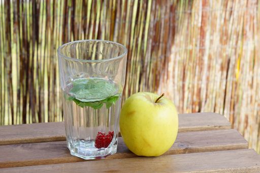 A closeup shot of a glass of water with mint and raspberry with an apple next to it