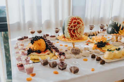 A beautifully arranged dessert table with carved fruits, various desserts, and elegant glassware at a sunny event.