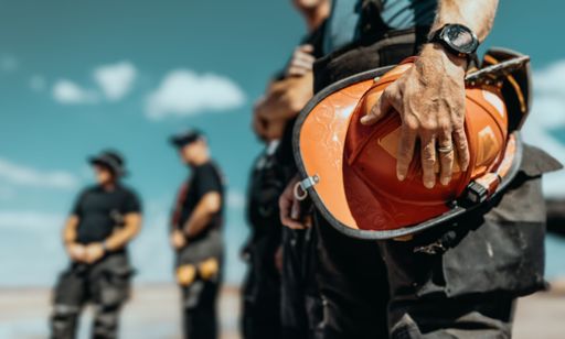 Close-up of a construction worker holding a hard hat with colleagues in the background under a clear sky.