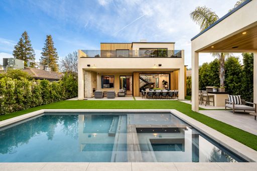 Modern luxury house with a swimming pool, outdoor seating, and lush greenery under a clear blue sky.