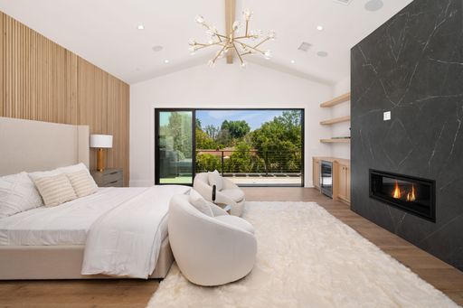 Modern bedroom with a cozy fireplace, stylish decor, and a large window overlooking greenery.