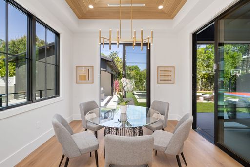 Modern dining area with a glass table and gray chairs, featuring large windows and a view of a backyard with greenery.