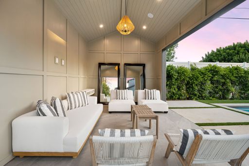 Modern outdoor seating area with white furniture and striped pillows, adjacent to a pool and lush green hedges at sunset.