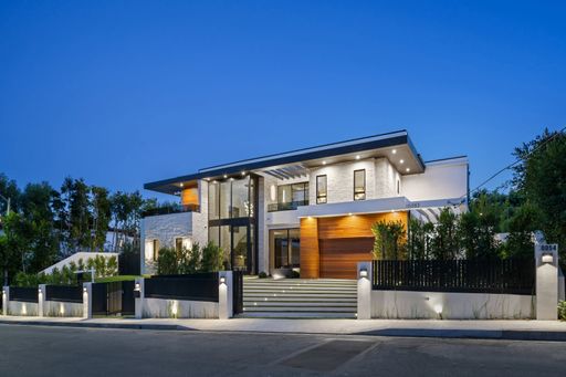 Modern luxury house exterior with large windows, illuminated at dusk, surrounded by greenery.