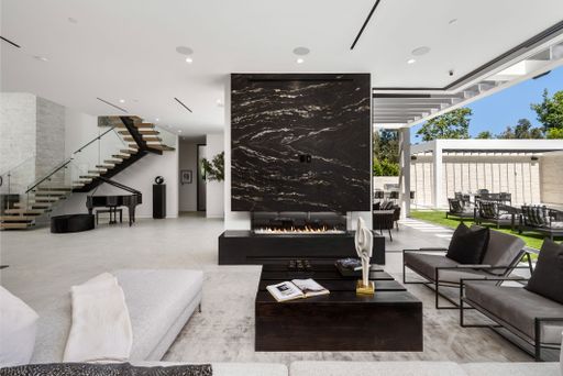 A modern living room interior with a black marble fireplace, open floor plan, and outdoor seating area visible through large windows.